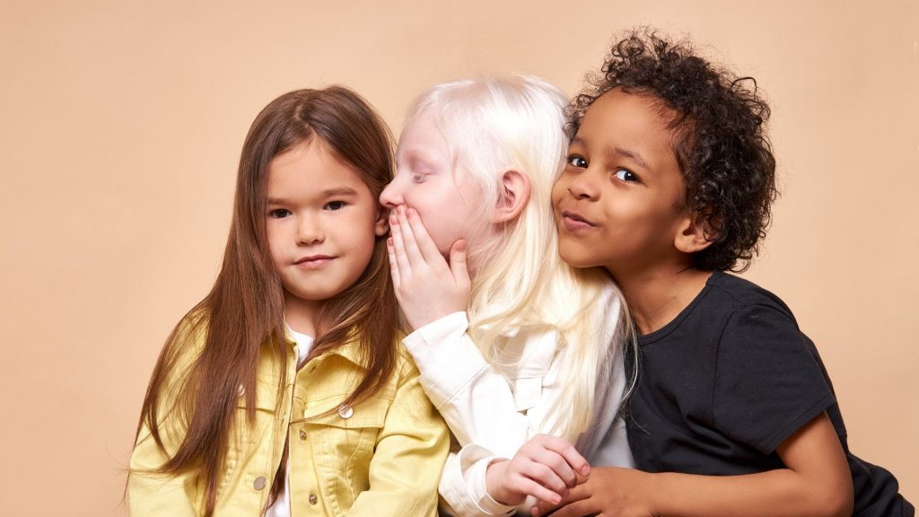 Trois enfants se passent des mots à l'oreille dans un décor uni rose